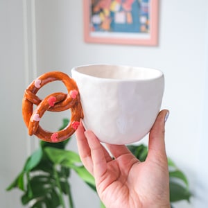 Puede incluir: Taza de cerámica blanca con un asa en forma de pretzel decorada con chispas rosas. La taza está siendo sostenida por una mano.