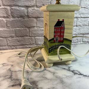 May include: A square wooden table lamp base with a cream-colored finish. The front panel features a hand-painted scene of a tall red house with a black roof, situated behind a black picket fence on a green grassy hill. A gold metal socket sits on top.