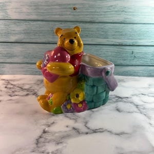 May include: A ceramic Winnie the Pooh planter. The yellow bear is holding a pink pot of honey and is next to a green and purple planter. The planter is decorated with flowers and has a white interior.