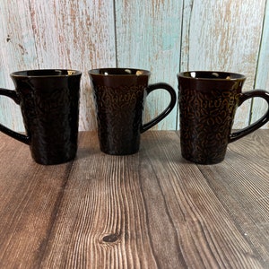 May include: Three brown ceramic coffee mugs with a coffee bean design. The mugs have a textured surface and a handle.
