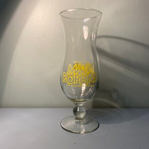 May include: A clear glass hurricane style glass with a gold inscription that reads "Disney's Mary Poppins The Broadway Musical".