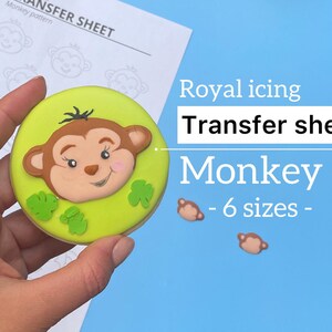 May include: A green cookie decorated with a brown monkey face and green leaves. The cookie is being held by a hand. The background is blue with the text "Royal icing Transfer sheets Monkey - 6 sizes - "