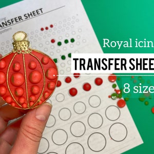 May include: A red and gold Christmas ornament cookie decorated with royal icing. The cookie is being held by a hand. The background shows a white sheet with a circle pattern and the text "Royal icing TRANSFER SHEET Circle pattern".