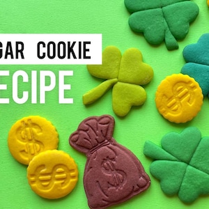 May include: A green background with the text "SUGAR COOKIE RECIPE" in white.  There are several sugar cookies in the shape of four-leaf clovers, money bags, and coins with dollar signs.