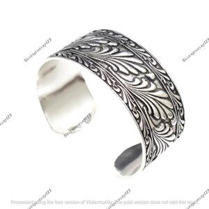 May include: A silver cuff bracelet with an ornate floral design. The open cuff has a detailed, embossed pattern across the top, with a smooth, polished interior. The bracelet is a wide style, suitable as a statement piece. The width is approximately 2.5 cm.