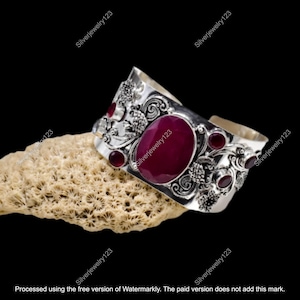 May include: A silver cuff bracelet with an oval, deep red gemstone at the center and smaller red gemstones. The bracelet has an ornate floral design. The bracelet is displayed on a beige, textured surface against a black background.