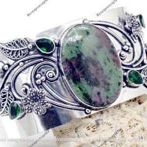 May include: A wide silver cuff bracelet featuring an oval-shaped, green and maroon stone at the center. The bracelet is adorned with green gemstones and floral and leaf designs. The jewelry is set on a textured surface.