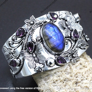 May include: A silver cuff bracelet with a central oval blue labradorite stone, complemented by smaller purple gemstones and floral and butterfly motifs. The bracelet showcases an intricate, detailed design.