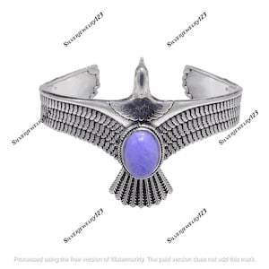 Pode incluir: Pulseira de punho prateada com um design de pássaro estilizado. O pássaro tem asas abertas e uma pedra ovalada roxa clara no centro. A pulseira apresenta padrões detalhados de penas e asas.
