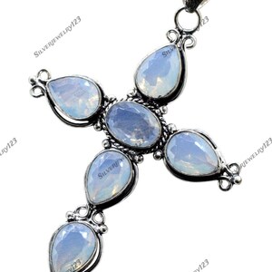 Puede incluir: Colgante plateado en forma de cruz con cinco piedras preciosas azul claro, de forma ovalada y de pera. El colgante tiene una argolla en la parte superior para una cadena y detalles decorativos plateados alrededor de las piedras.