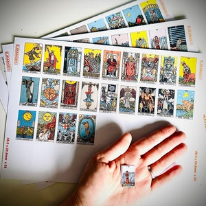 May include: A set of tarot cards with colourful illustrations and gold accents. The cards are laid out on a white surface, with a single card held in a hand. The cards measure 48.9 x 29.6 mm.