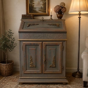 Op de afbeelding: Een grijze secretaire met een neerklapbaar schrijfblad en twee onderste kastdeuren. Het bureau is voorzien van goudkleurige decoratieve hoekaccenten, een centrale metalen handgreep en een rechthoekig bovengedeelte met een scharnierend deksel.