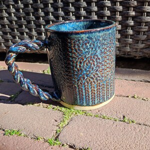 May include: A blue ceramic mug with a rope-like handle and a textured design. The mug has a white rim and base.
