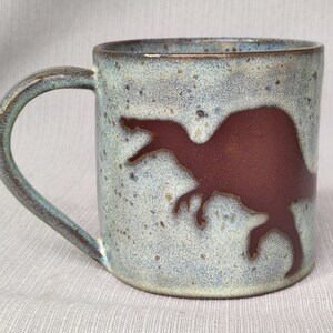 Op de afbeelding: Een keramische mok met een gespikkeld lichtblauw glazuur en een bruin dinosaurus silhouet.
