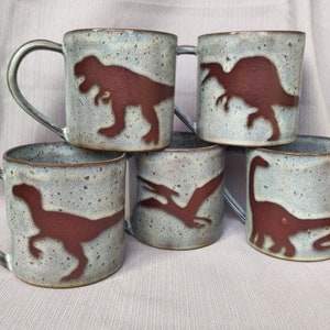 May include: Set of five ceramic mugs with dinosaur silhouettes. Each mug features a different dinosaur in a reddish-brown color against a speckled light blue and gray background.