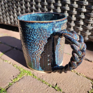 May include: A blue ceramic mug with a braided handle. The mug has a textured surface with a dark blue and brown glaze.