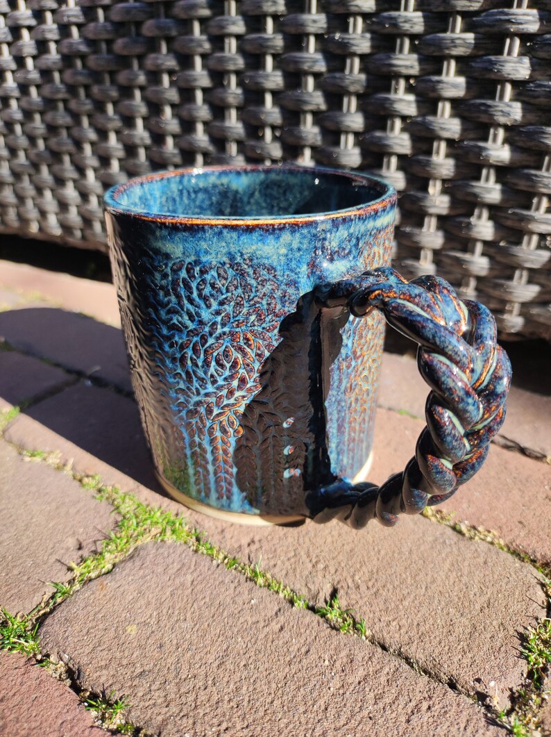 May include: A blue ceramic mug with a braided handle. The mug has a textured surface with a dark blue and brown glaze.