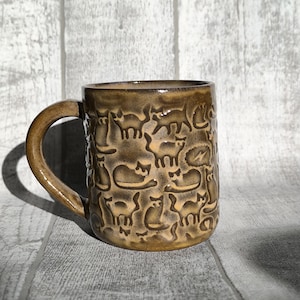 May include: A brown ceramic mug with a pattern of cats all over it. The mug has a handle and is sitting on a white wooden surface.