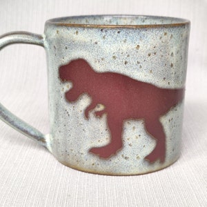 Può includere: Una tazza in ceramica con una glassa blu chiaro macchiata e un bordo marrone. La tazza ha una silhouette di dinosauro rossa sul davanti.