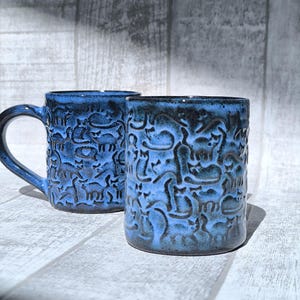 May include: Two blue ceramic mugs with a textured surface and a cat design. One mug has a handle, while the other is handle-less. The mugs are a deep blue color with darker accents, and are set against a light-colored background.