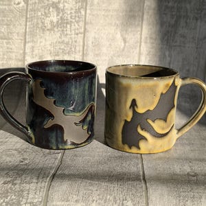 Op de afbeelding: Twee keramische mokken met een zwart-wit silhouetontwerp. De ene mok is donkerblauw en groen, de andere is geel en bruin.