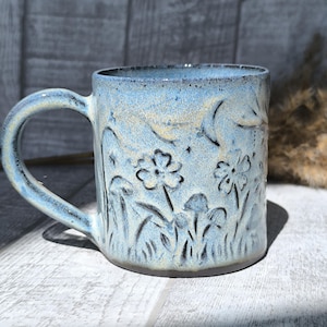 Peut inclure: Une tasse en céramique avec un émail bleu clair présentant un motif en relief de fleurs sauvages et de champignons. La surface présente des nuances jaunes subtiles et des contours sombres définis autour des motifs botaniques, avec une anse.