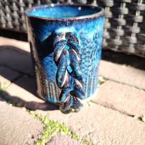 May include: A blue ceramic mug with a unique, braided handle. The mug has a textured, speckled glaze with a dark blue and brown color scheme.
