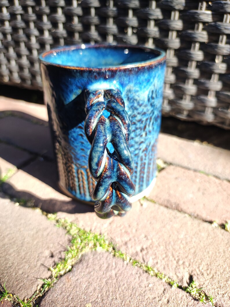 May include: A blue ceramic mug with a unique, braided handle. The mug has a textured, speckled glaze with a dark blue and brown color scheme.