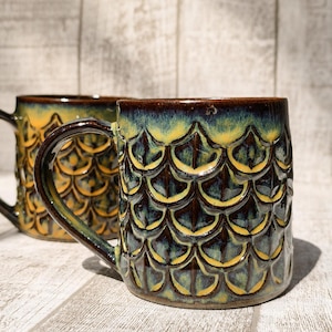 Può includere: Due tazze in ceramica con un motivo strutturato a forma di scaglie. Le tazze presentano un bordo e un manico marrone scuro, con un gradiente di tonalità gialle, verdi e blu all'esterno. Le tazze sono su uno sfondo di legno chiaro.