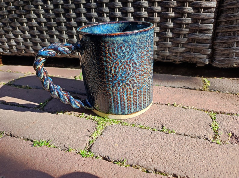 May include: A blue ceramic mug with a rope-like handle and a textured design. The mug has a white rim and base.