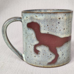 Op de afbeelding: Een keramische mok met een gespikkeld lichtblauw glazuur en een bruin dinosaurus silhouet.