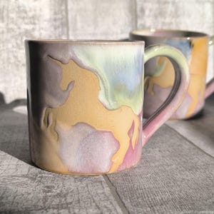 Könnte beinhalten: Eine Keramiktasse mit einem pastellfarbenen Einhorn-Design. Die Tasse hat eine hellviolette, gelbe und rosa Farbgebung mit einer geformten Einhorn-Silhouette. Der Griff ist rosa. Eine weitere Tasse ist im Hintergrund zu sehen.