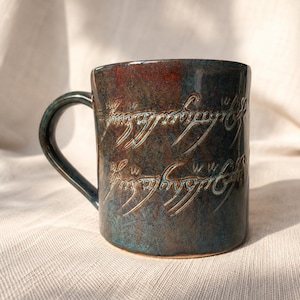Könnte beinhalten: Eine Keramiktasse mit dunkelblau-grüner und brauner Glasur. Die Tasse hat eine geprägte Inschrift in einer stilisierten Schriftart. Der Griff hat eine passende dunkelblau-grüne Farbe. Die Tasse steht auf einer weißen Oberfläche.