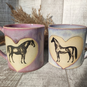 Pode incluir: Duas canecas de cerâmica com um design em forma de coração que apresenta uma silhueta preta de um cavalo. As canecas são em tons de roxo e azul.