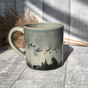May include: A grey ceramic mug with a black silhouette of a forest and birds flying overhead. The mug has a handle and is sitting on a wooden surface.