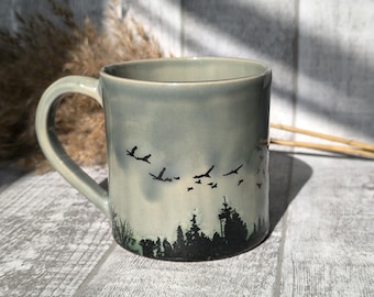 May include: A gray ceramic mug with a black silhouette of a forest and birds flying overhead. The mug has a handle and is sitting on a wooden surface.