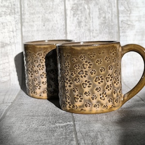 May include: Two brown ceramic mugs with a paw print design. The mugs are on a white wooden surface.
