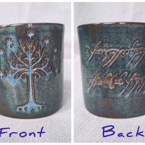 May include: A ceramic mug with a blue-green glaze. The front of the mug features a brown tree with white stars. The back of the mug features the inscription "One Ring to rule them all, One Ring to find them, One Ring to bring them all and in the darkness bind them." in Elvish script.