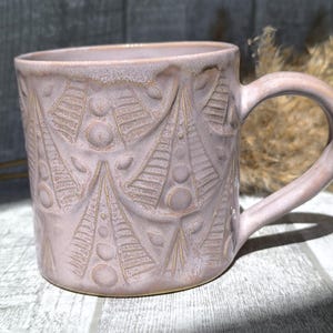 Pode incluir: Uma caneca de cerâmica rosa claro com um padrão em relevo de formas triangulares estilizadas e pontos circulares. A caneca apresenta um acabamento liso e brilhante, uma asa curva no lado direito e um corpo cilíndrico.