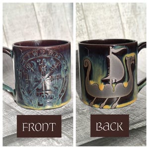 May include: A ceramic mug with a dark brown glaze and a teal and yellow underglaze. The front features a compass rose design, while the back displays a Viking ship. The mug has a handle and is labeled "FRONT" and "BACK".