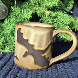 May include: A handcrafted ceramic mug with a dark brown pattern on a tan and brown surface. The mug features a curved handle and is set against a Christmas tree background.