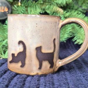 Puede incluir: Una taza de cerámica con exterior beige y asa marrón. La taza presenta dos siluetas de gatos marrón oscuro. La taza está sobre un fondo borroso de un árbol de Navidad y una superficie de punto azul.