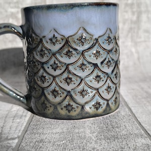 May include: Ceramic mug with a light blue glaze and a textured, scale-like pattern in shades of blue and brown. The mug features a handle and a detailed design, perfect for enjoying hot beverages.