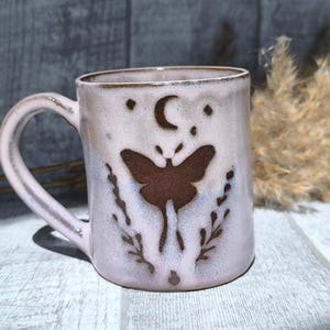 Peut inclure: Une tasse en céramique avec une finition émaillée rose clair et blanche, ornée d'un motif de papillon de nuit en relief marron foncé au centre. Au-dessus, un croissant de lune et des étoiles, avec des branches feuillues de chaque côté.