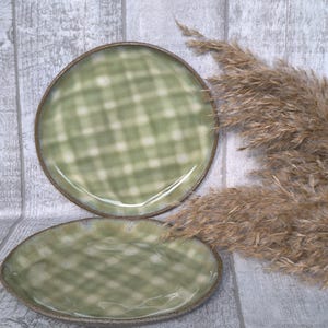 Pode incluir: Dois pratos redondos de cerâmica com um padrão suave de xadrez em verde sálvia e branco. Os pratos têm um acabamento brilhante e uma borda castanha escura texturizada. O design é consistente em ambas as peças, exibindo uma aparência subtil e artesanal com bordas lisas e ligeiramente irregulares.