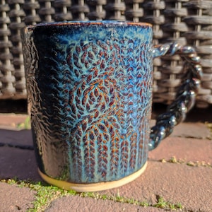 May include: A blue ceramic mug with a textured, leafy design and a twisted handle. The mug has a dark blue glaze with hints of brown and white.