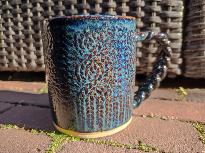 May include: A blue ceramic mug with a textured, leafy design and a twisted handle. The mug has a dark blue glaze with hints of brown and white.