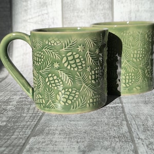 May include: Two green ceramic mugs with an embossed pattern of pinecones, needles, and stars. The mugs have a curved handle and a glossy finish. The mugs are displayed on a grey wooden surface.