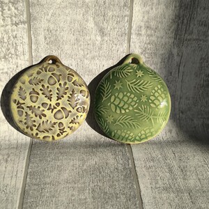 May include: Two ceramic ornaments with a winter theme. One ornament is green with pine needles and pinecones. The other ornament is yellow with acorns and oak leaves.