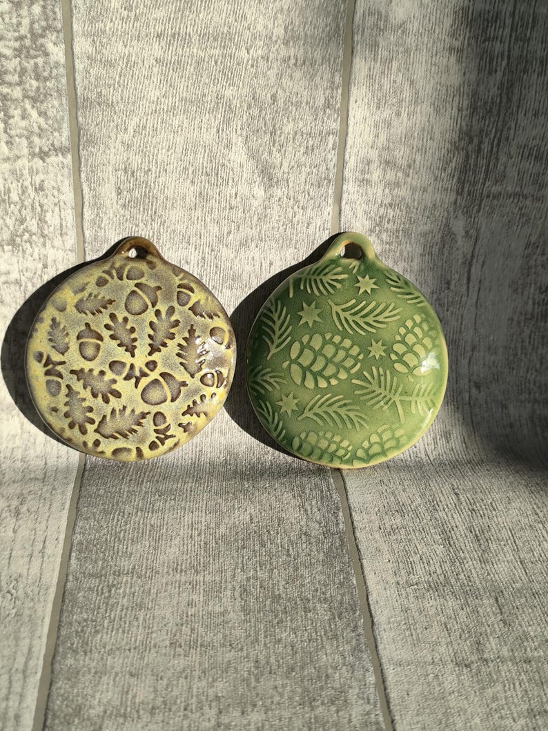 May include: Two ceramic ornaments with a winter theme. One ornament is green with pine needles and pinecones. The other ornament is yellow with acorns and oak leaves.